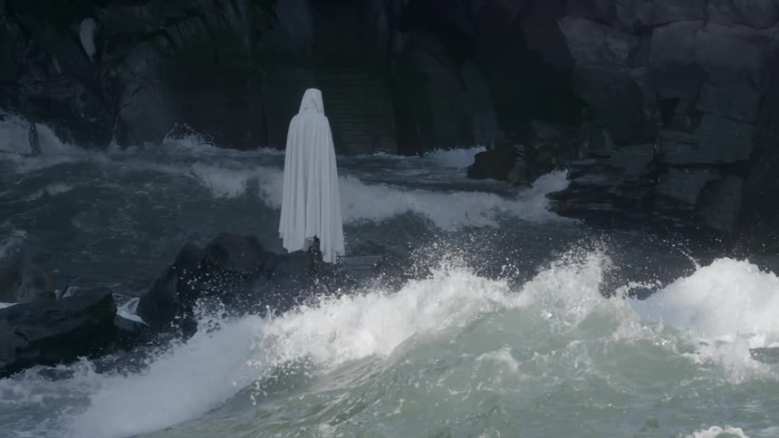 A person wearing white robes stands on rocks in the center of a rapid
