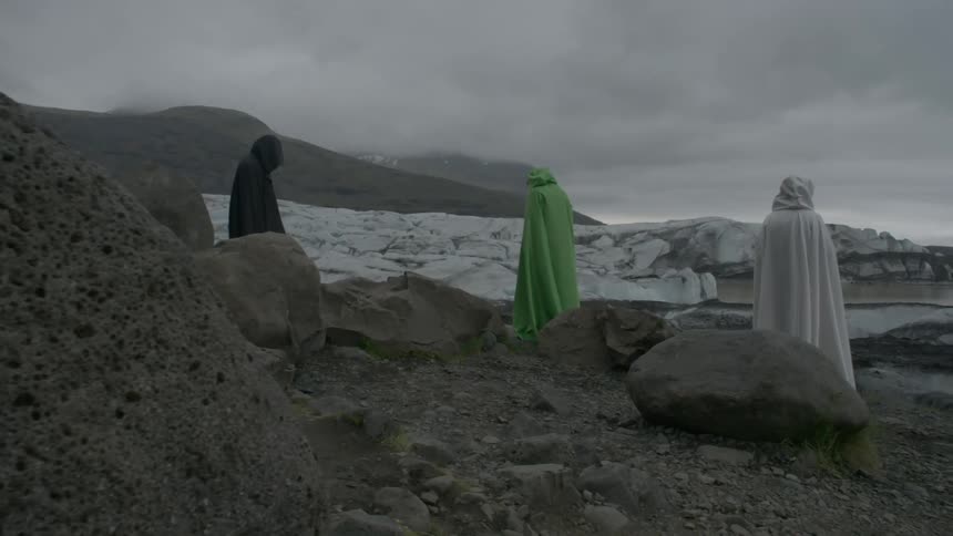 Three persons wearing white, black or green cloaks stand among rocks