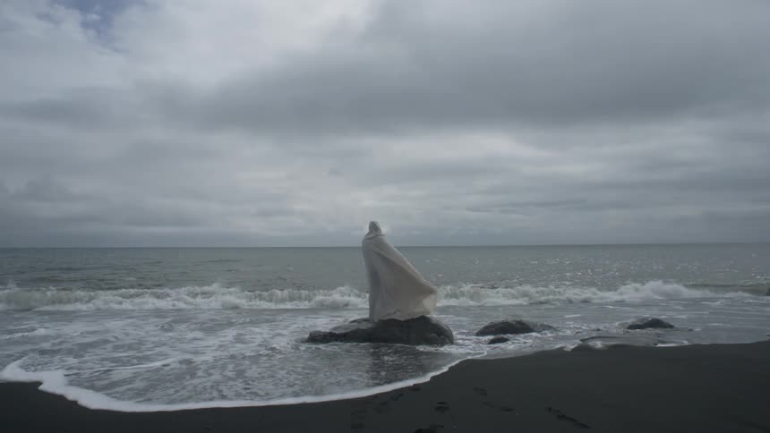 A person wearing white robes is standing on a rock by the shore