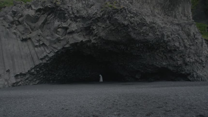 A person wearing white robes stands inside a dark cavern in the distance
