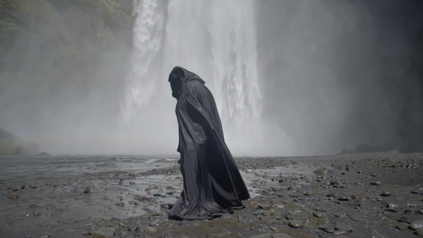 A person wearing dark robes is standing at the foot of a waterfall