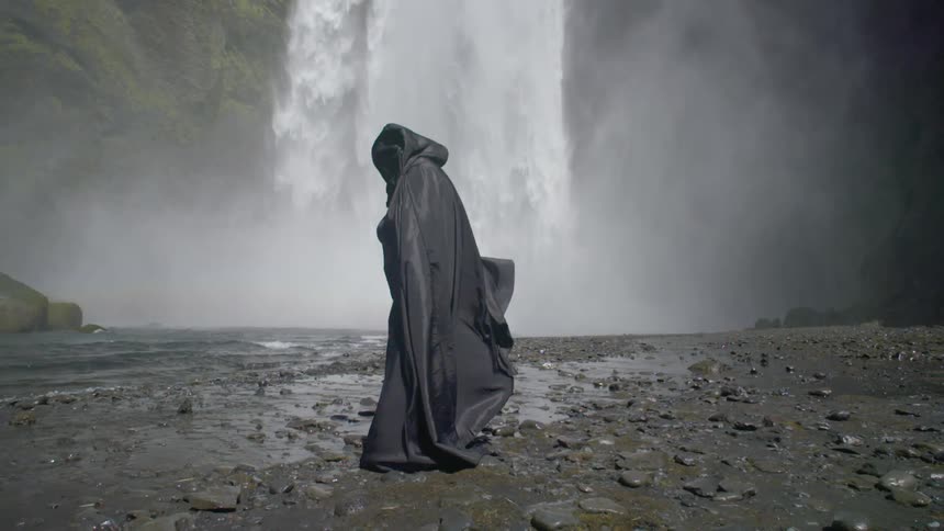 A person wearing black robes is standing near the bottom of a waterfall