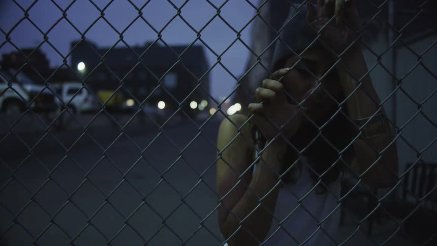 Woman standing near wired fence on city street at sunset