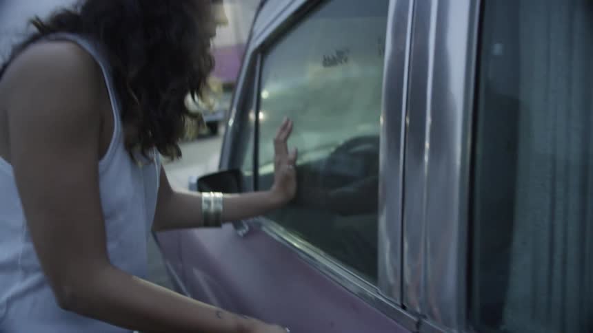 A woman pulls on a car door handle and looks inside the driver side window