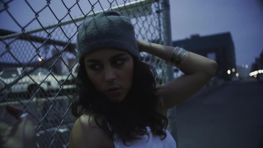 Two women stand against a chain link fence near a parking lot in a city neighborhood at night