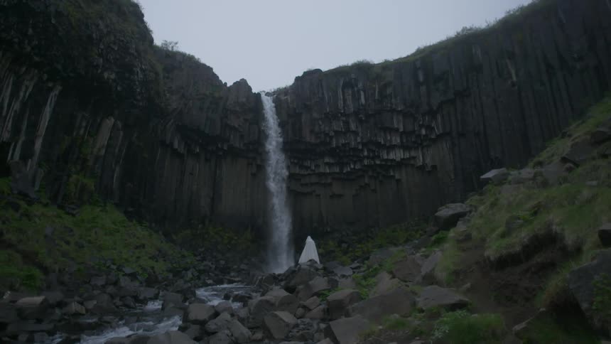 A person wearing a white robe is standing at the foot of a waterfall