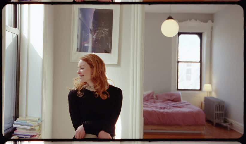 A woman sits in her bright sunny apartment smiling and laughing then leaning back to look out the window