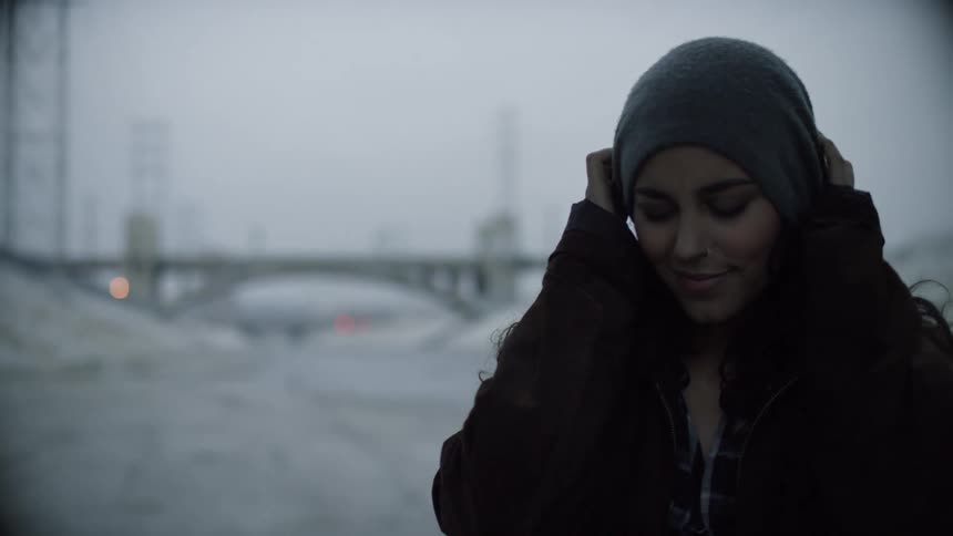 A woman tugs on a beanie on her head outside near a bridge