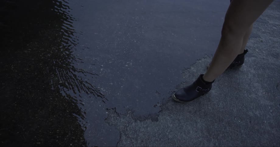 A woman wearing boots with bare legs walks to the edge of a puddle on a street
