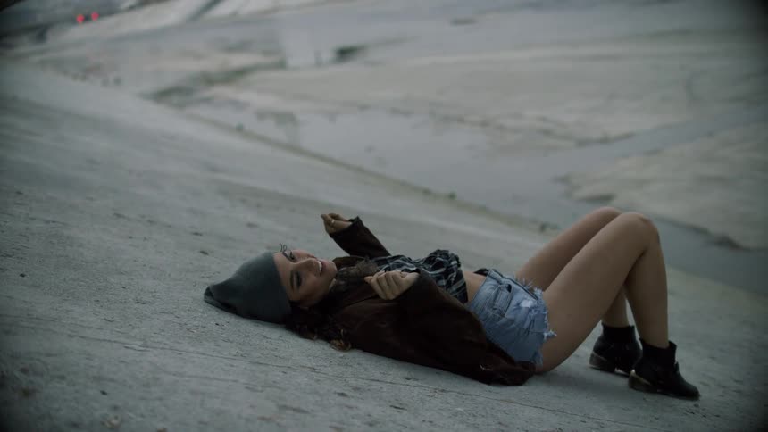 A teenager lies on a concrete embankment snapping her fingers