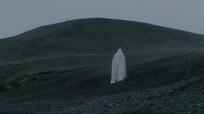 A person wearing white robes is standing on some hills on a cloudy day