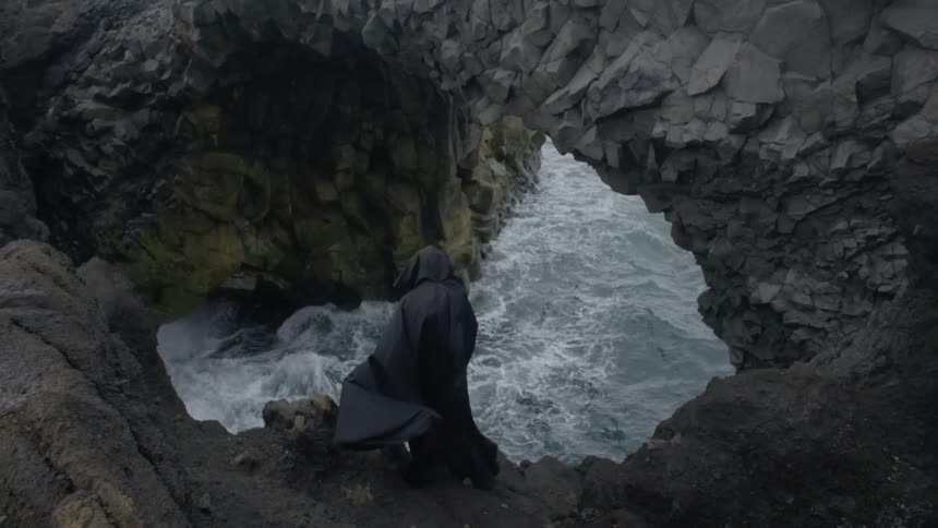 A black caped figure stands on a rocky ledge looking down at the water