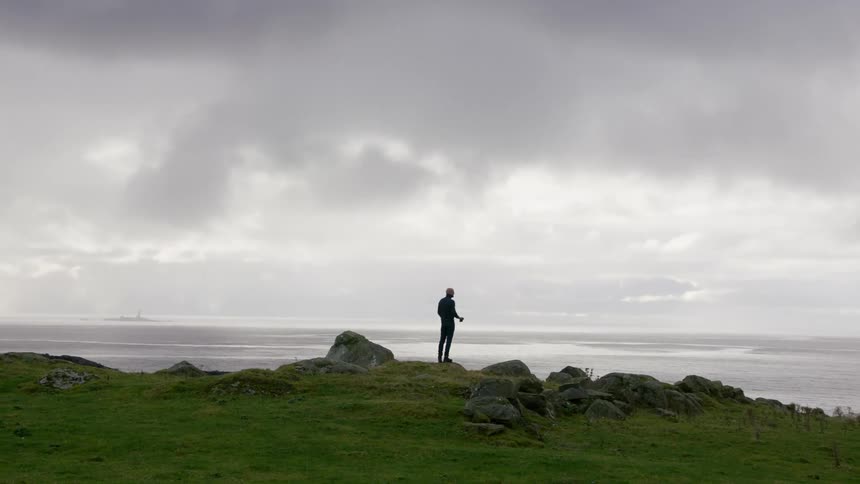 Man standing at the edge of a cliff looks at the view through binoculars