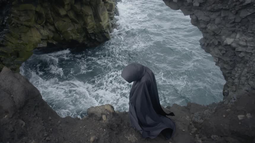 A person cloaked in a black hooded robe stands on a rock ledge over rushing water