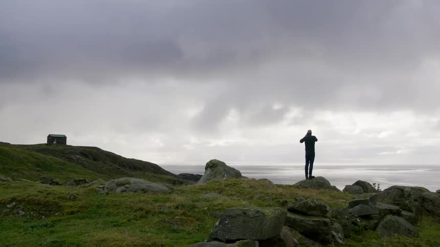 A person standing atop a rock on a hilly cliff taking photographs of the ocean
