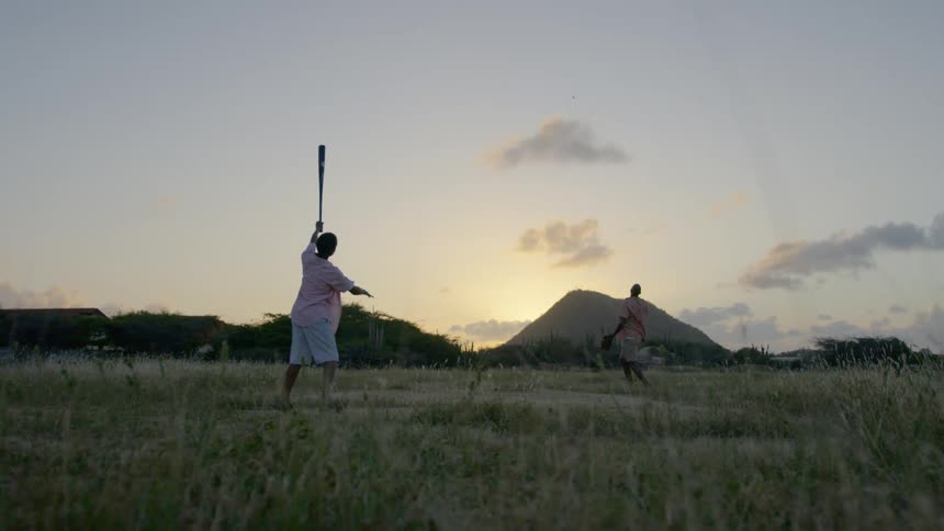 Sun shines on horizon as boy and man play baseball in field near buildings
