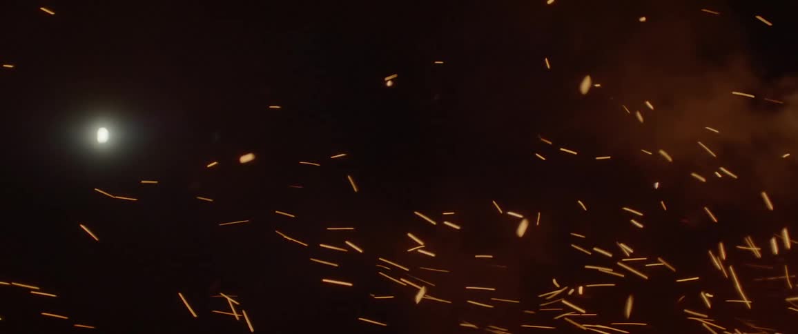 Sparks embers and smoke rise up from a fire while the moon glows in the night sky