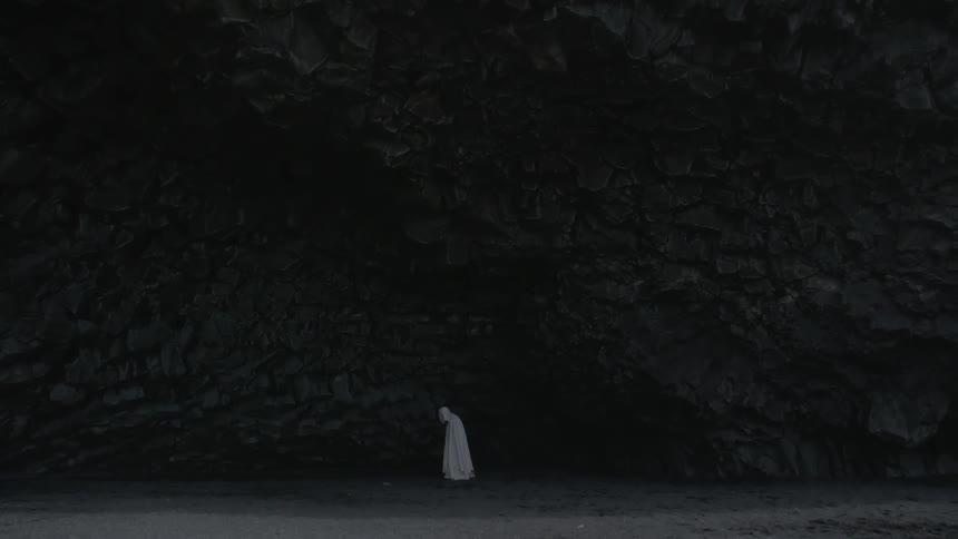 A white caped figure stands at the base of a rocky cliff