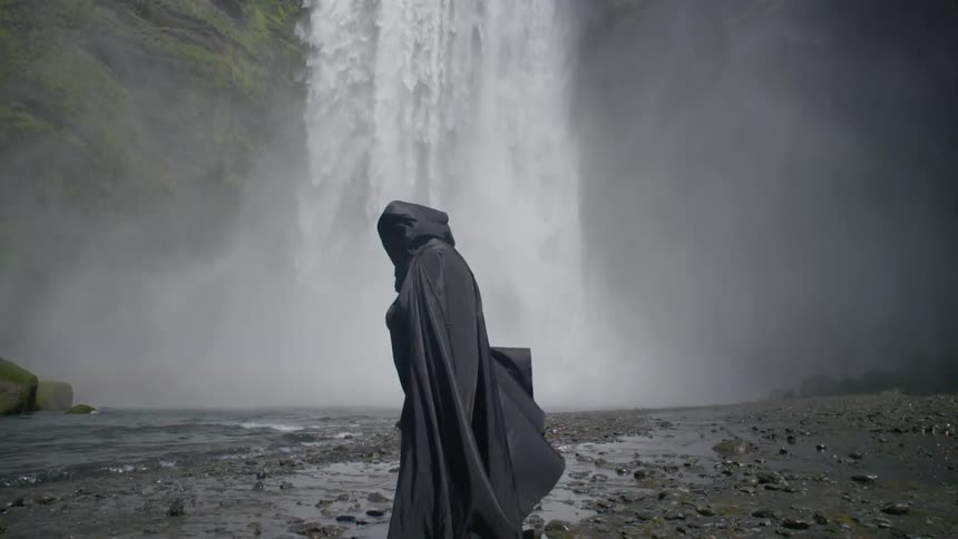 A person wearing black robes is standing at the foot of a waterfall