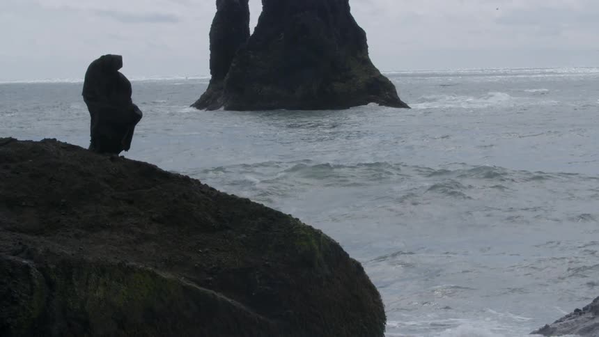 A person wearing black robes is standing on some rocks by the shore