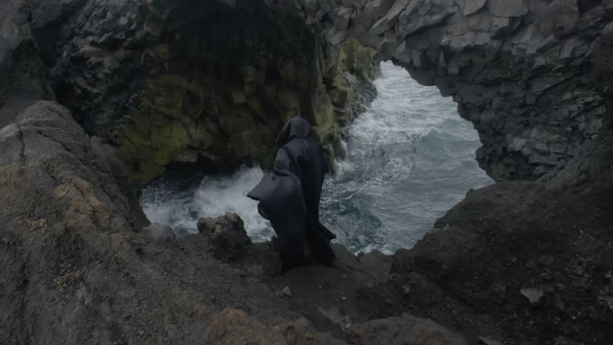 A person sheathed in black fabric stands at the water's edge on a rocky formation