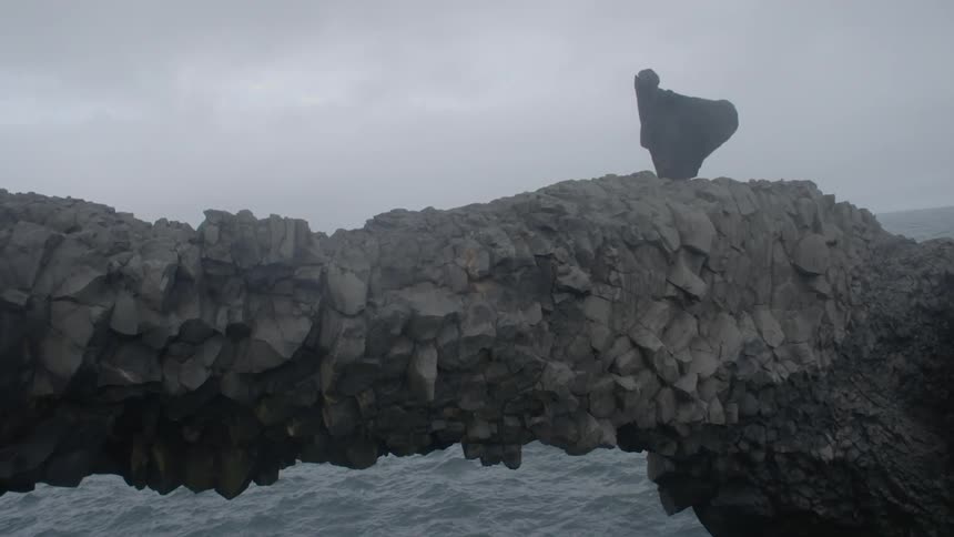 A person wearing black robes is standing on some rocks by the water
