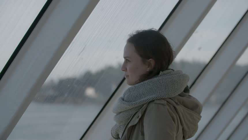 A woman is standing near a large window looking over water