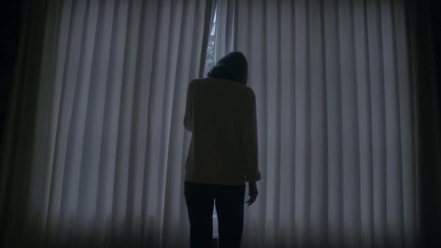 A woman walks over to a window and peaks through the blind