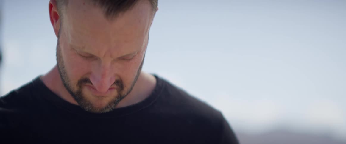 A white man with a beard stands outdoors and puts his head down sadly