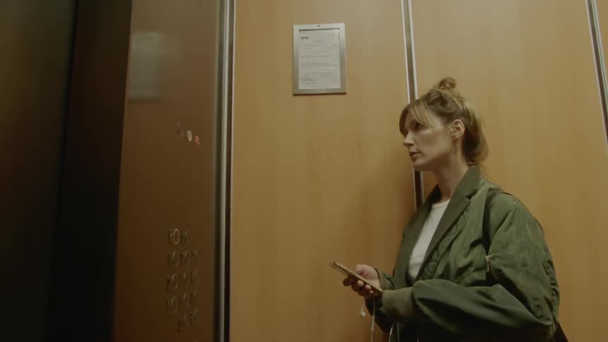 A woman stands in an elevator using a smartphone before grinning as the elevator door opens