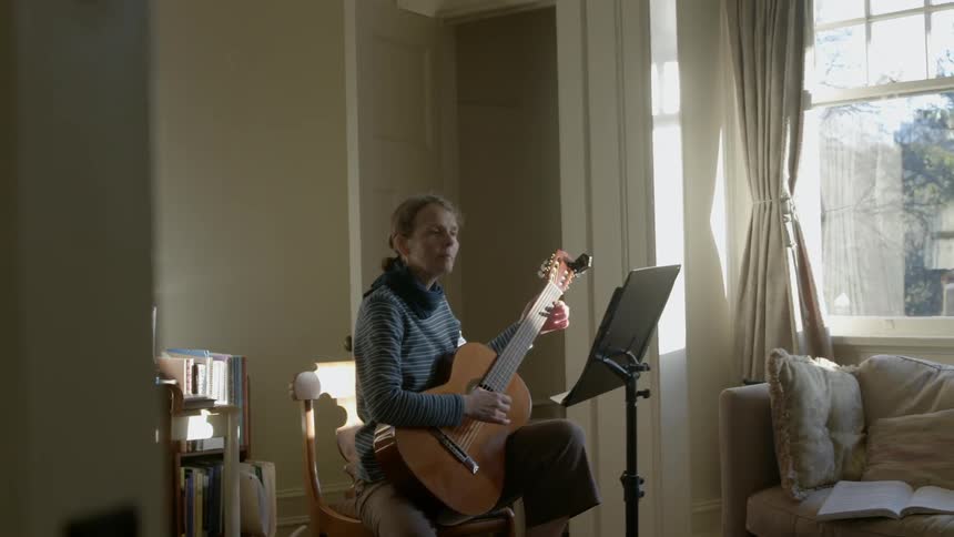 A woman plays guitar while sitting in a chair next to a window