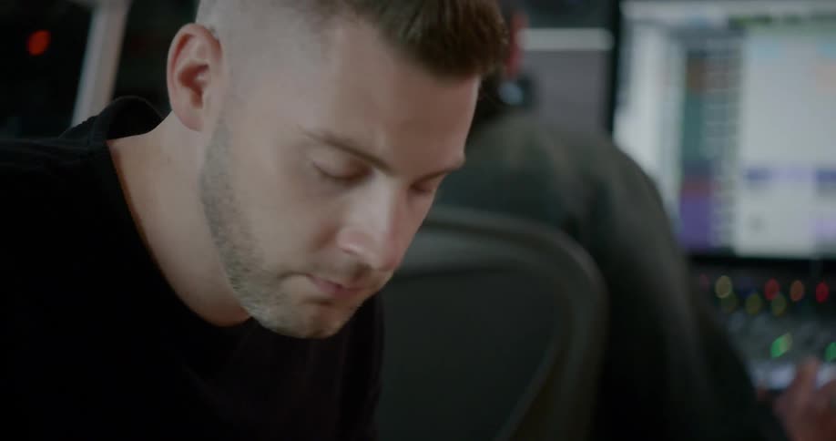 A man sitting down listening to music and shaking his head while another man is in the background touching controllers