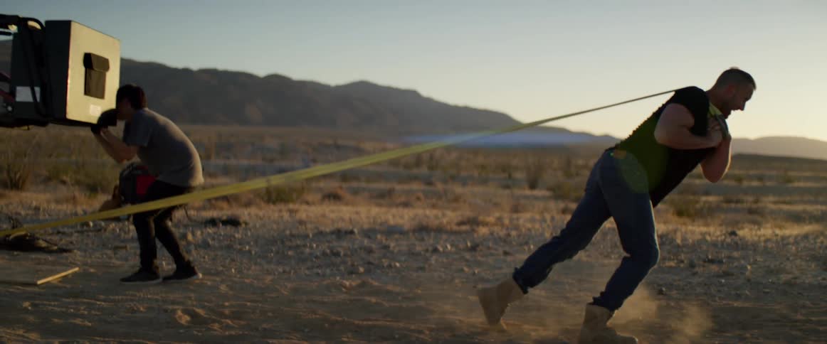A man uses rope to pull a crane as people film the scene in a field near a mountain range