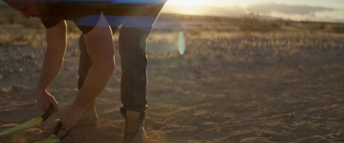 A man shakes out his arms then bends to pick up straps with handles and pull them over his shoulder in the desert