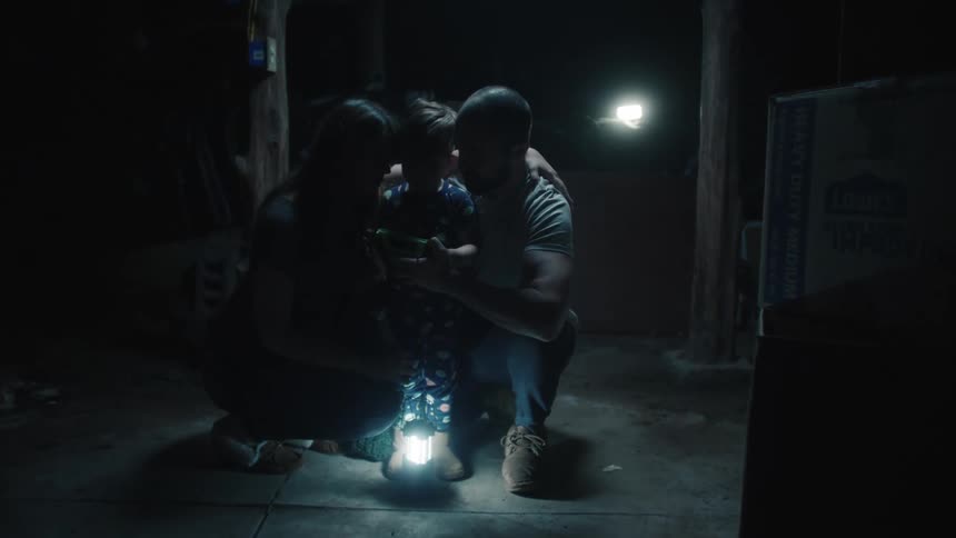 Parents are crouched down on either side of their son as they keep him distracted with a toy in a storm cellar