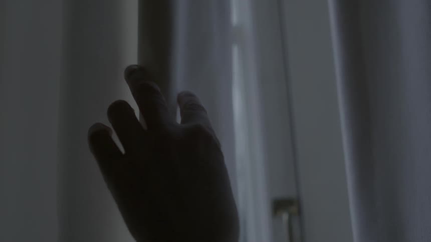 A hand slowly closes a curtain by a window