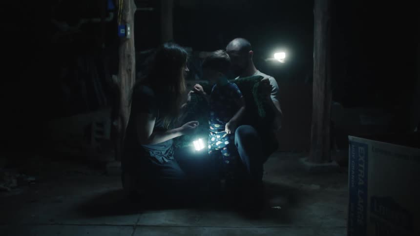 A mother and father comfort their son as they stand in a dark empty room with only two handheld lamps for light