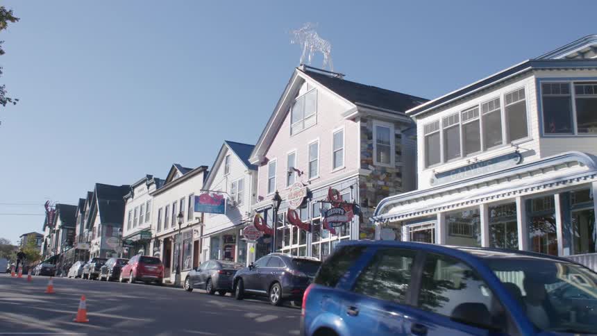 People ride bikes on a road past quaint shops on a sunny day