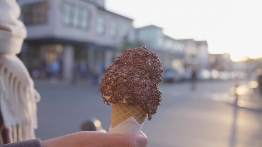 Woman holds an ice cream cone and raises it to her mouth as she stands near the city street