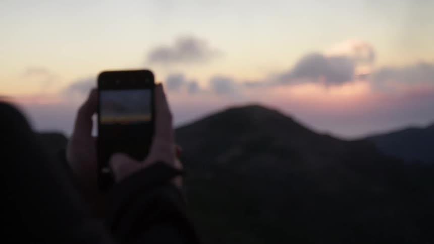 A man stands outdoors and photographs a mountain range