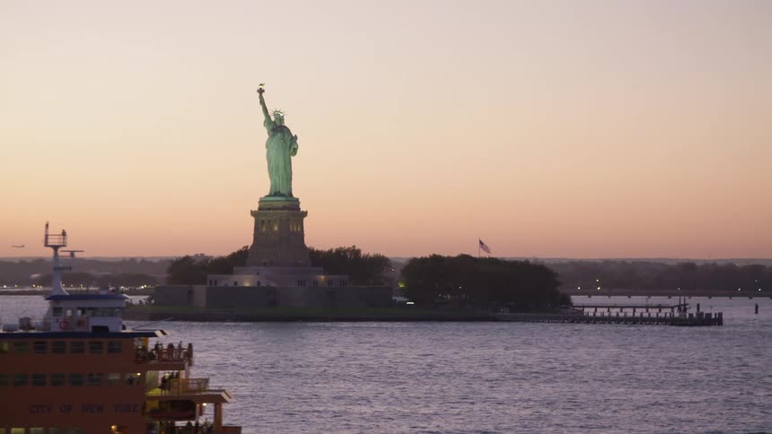 Passenger laden ferry passes near statue of liberty backed by orange sky at the horizon