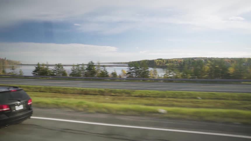 Car drives along highway past forest covered shore and water under cloudy sky