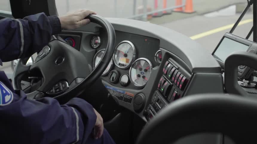 A bus driver wearing a uniform jacket with a patch on it turns the wheel