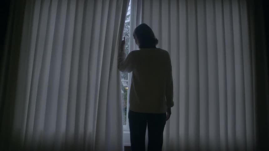 A woman walks over to window and peeks past the blinds