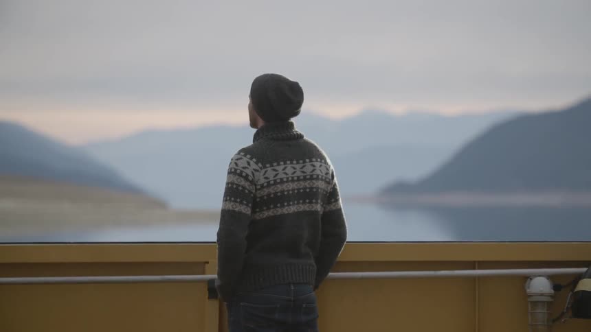 A man in a sweater and hat looks out towards a mountain lake
