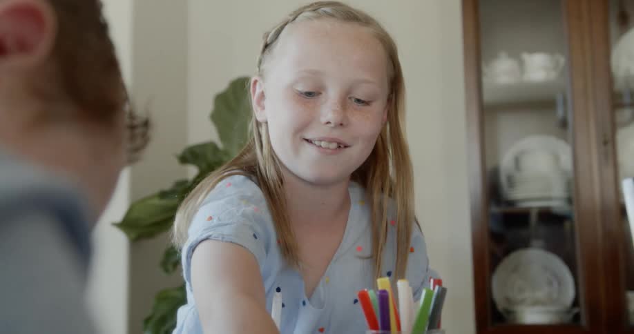 Two children sit at a dining room table and work with pens