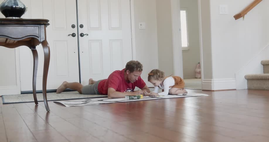 A dad lies on the floor with his daughter coloring on a large sheet of paper in their home