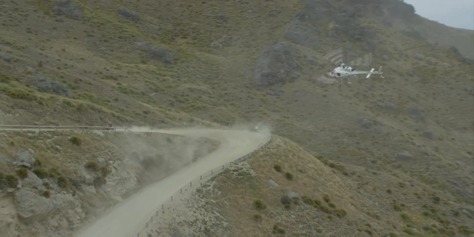 A helicopter hovers in the air as a vehicle races around a dirt road in the desert