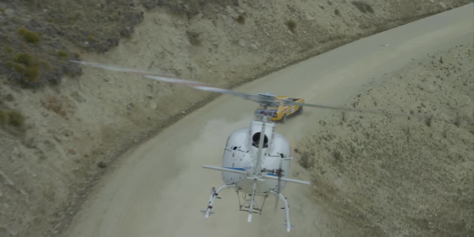 White helicopter closely follows yellow and black car driving fast along dirt road