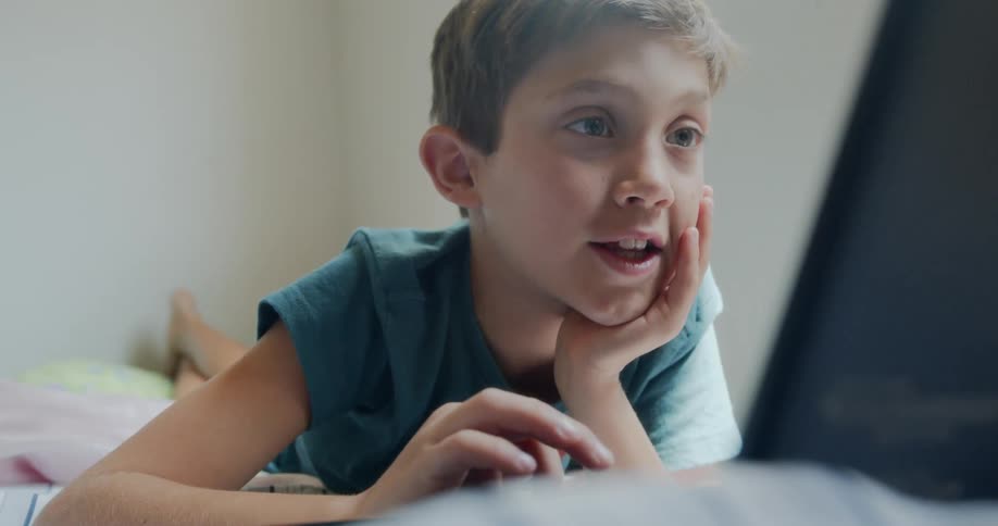 A young boy lays on his bed and plays with a laptop
